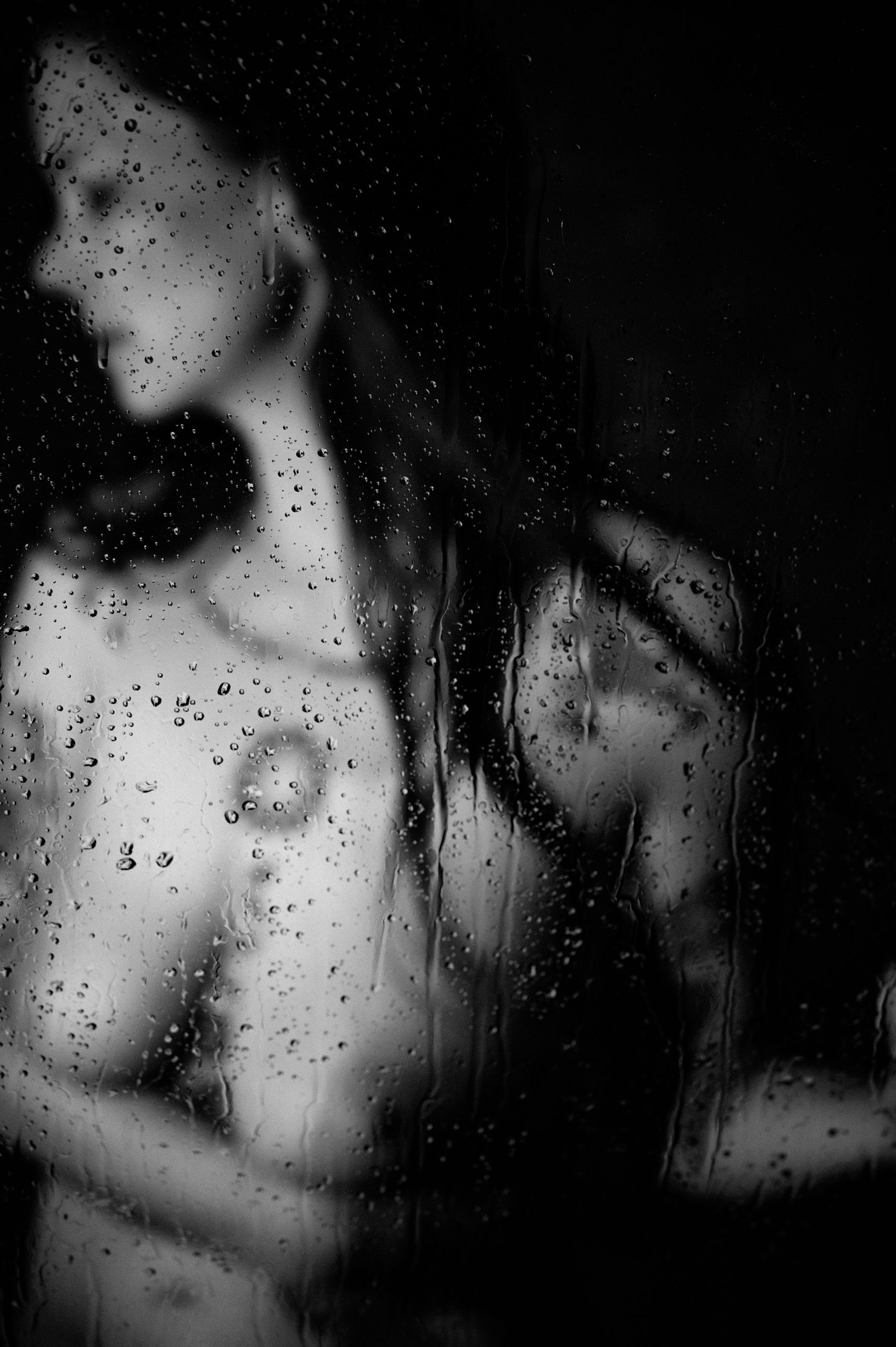 image of a woman in a boudoir session, she is in the shower. The camera is focused on the water drops on the shower door so she is slightly out of focus but you can still make out her tattoos and see that her face is turned to the left away from the camera.