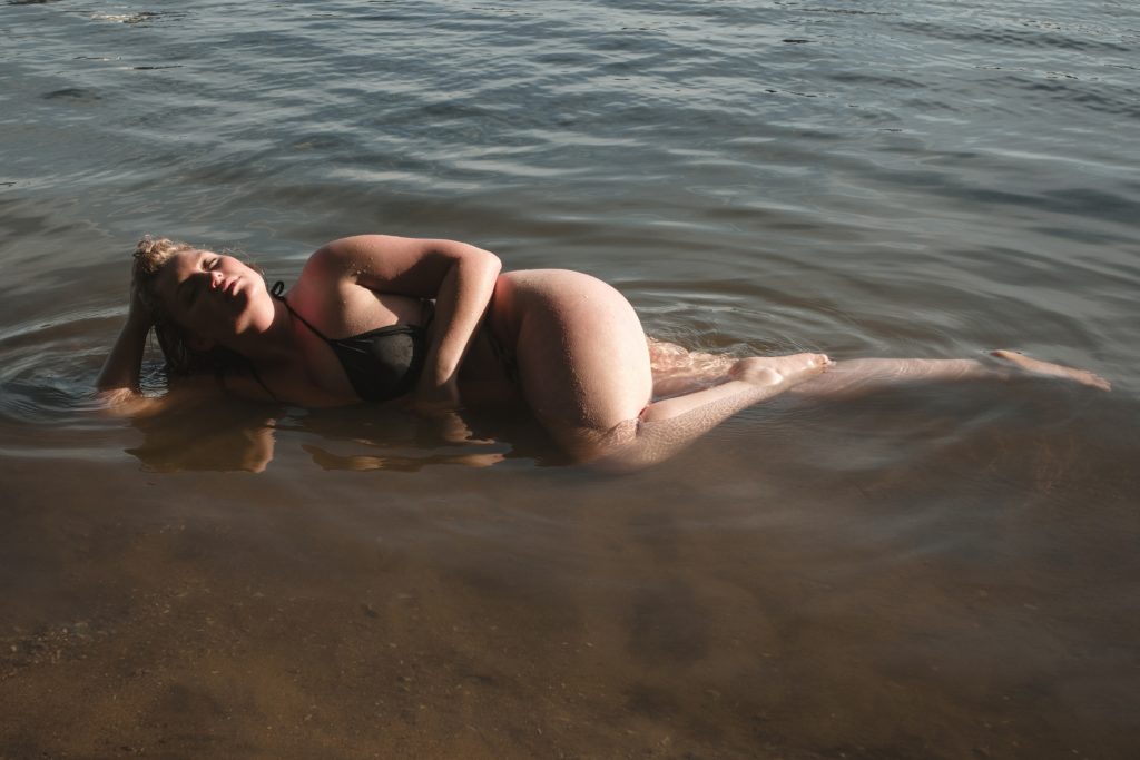 an outdoor boudoir session of a woman laying on her side in shallow river water in a black bikini, her face is turned up towards the harsh sunlight and her eyes are closed