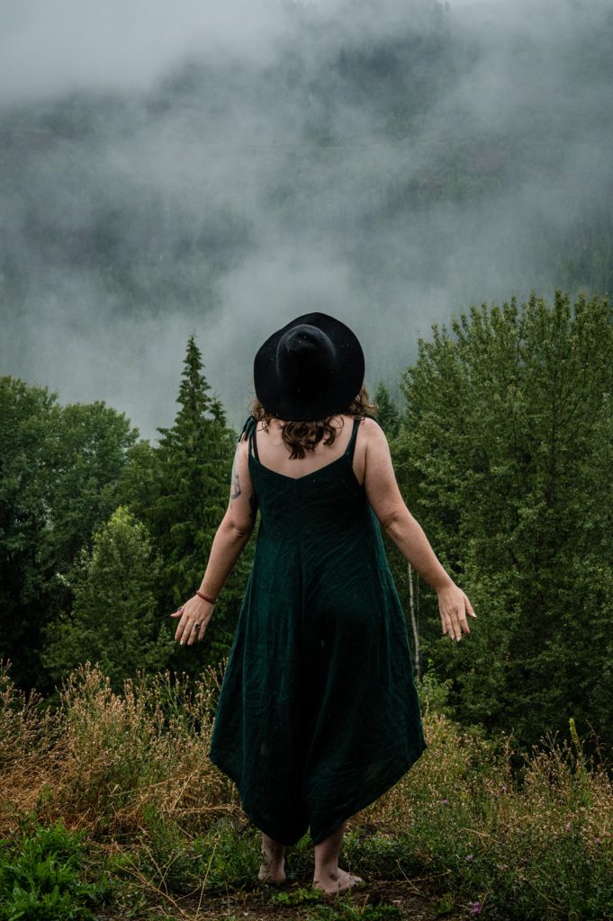 woman in a green romper standing in mountain pose with a black hat on, there is mist in the background in front of her and she is surrounded by green trees and forest