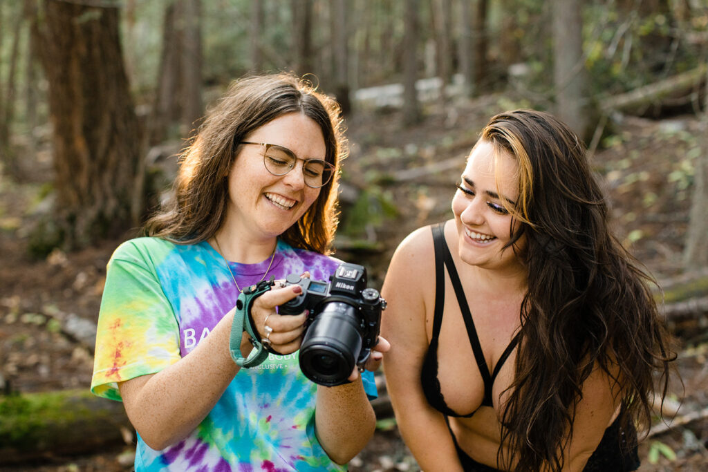 boudoir photographer Erika Broom shows her client the back of her camera during an empowerment photography session. They are both smiling and looking at the camera, the woods of BC can be seen behind them.